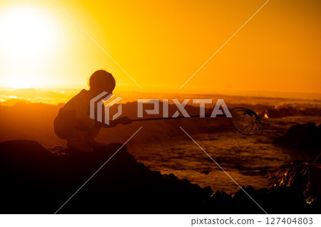 Sunset, silhouette and child on a rock at the beach playing or fishing with net in the water. Travel, seaside and girl kid in nature by the ocean in the evening to play or have fun while on vacation. 127404803