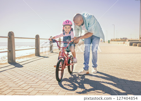 Father, child and bicycle with a girl learning to ride a bike on promenade by sea for fun, bonding and quality time on summer vacation. Man teaching his daughter or girl safety while cycling outdoor 127404854