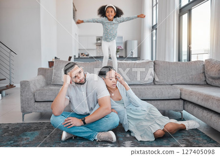 Tired, parents and child jump on sofa with exhausted mother and father sitting on floor in living room. Family, energy and young girl jumping on couch with frustrated, annoyed and upset mom and dad 127405098