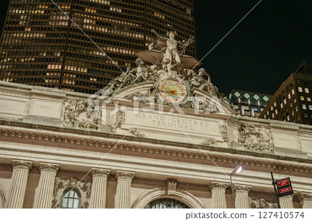 The famous Grand Central Terminal clock is illuminated above the main entrance, surrounded by classical statues. Tall skyscrapers tower behind the detailed architecture in Midtown Manhattan. 127410574