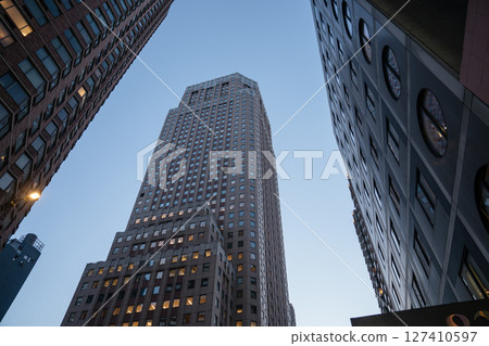 A towering skyscraper is framed by other high-rise buildings in New York City during early evening. The sky is clear as city lights begin to glow from office windows. A towering skyscraper is framed by other high-rise buildings in New York City during early evening. The sky is clear as city lights begin to glow from office windows. 127410597