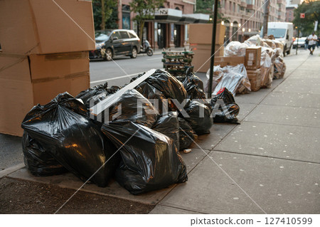 A pile of black garbage bags is stacked along a city sidewalk beside large cardboard boxes. The street scene shows an urban environment with buildings, cars, and early evening light. 127410599