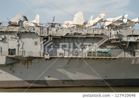 Multiple vintage Navy fighter jets are displayed on the upper deck of a docked aircraft carrier. Below, the steel hull shows staircases, rust stains, and architectural ship details above calm water. Multiple vintage Navy fighter jets are displayed on the upper deck of a docked aircraft carrier. Below, the steel hull shows staircases, rust stains, and architectural ship details above calm water. 127410646