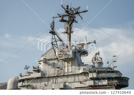The communication and radar tower of a military aircraft carrier rises high with multiple antennas and satellite dishes. The complex steel framework is silhouetted against a clear blue sky. 127410649