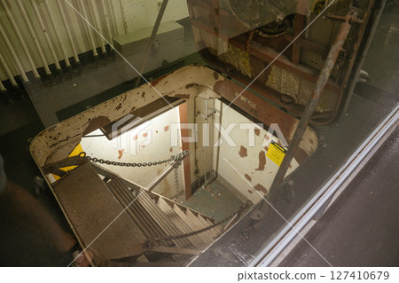 View of a steep metal ladder leading down a worn submarine hatch. Chipped paint, warning sign, and exposed pipes give an industrial feel. 127410679
