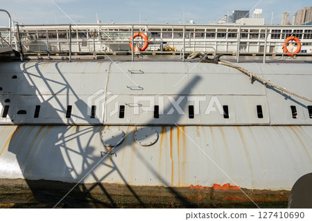 Detailed view of a grey military submarine hull with rust streaks, shadows of railing structures, and two access hatches. Dock equipment and lifebuoys are visible above the vessel. 127410690