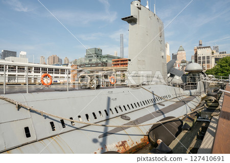Side view of a docked submarine with visible conning tower and ropes tied to a pier. The background features modern urban buildings and a clear sky. 127410691