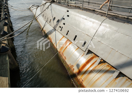 Detailed view of a submarine's hull showing streaks of rust and waterline marks. The vessel is docked at a wooden pier with ropes securing it above the water. 127410692