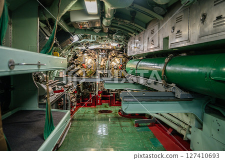 A navy submarine torpedo room shows twin torpedo tubes, a green torpedo secured in storage, and metal bunks along the wall. Overhead lighting illuminates control panels, piping, and equipment. 127410693