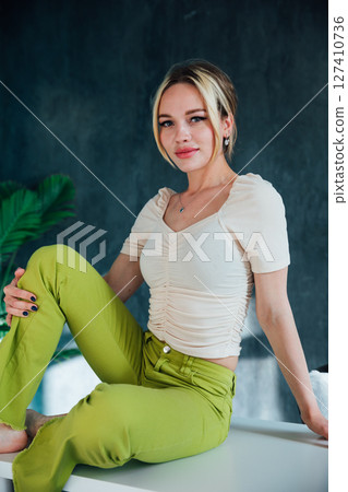 Beautiful blonde woman in green pants sitting on a white table Beautiful blonde woman in green pants sitting on a white table 127410736