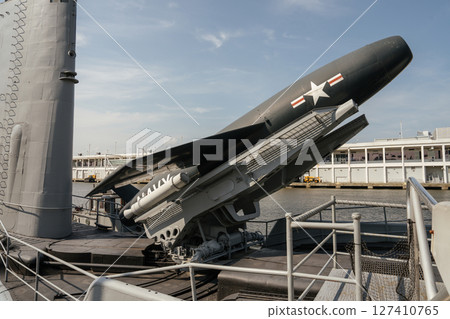 A large naval missile is displayed on the deck of a preserved U.S. Navy submarine at a waterfront museum. The submarine is docked near a city harbor under a clear blue sky. 127410765