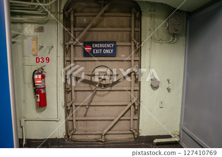 A secure emergency exit door with a wheel-lock mechanism is mounted on a ship or industrial wall. A red fire extinguisher is attached beside the hatch labeled. A secure emergency exit door with a wheel-lock mechanism is mounted on a ship or industrial wall. A red fire extinguisher is attached beside the hatch labeled. 127410769