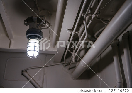 A glowing industrial-style ceiling lamp is mounted among painted metal pipes and conduit lines. The scene has a utilitarian and minimal aesthetic. 127410770