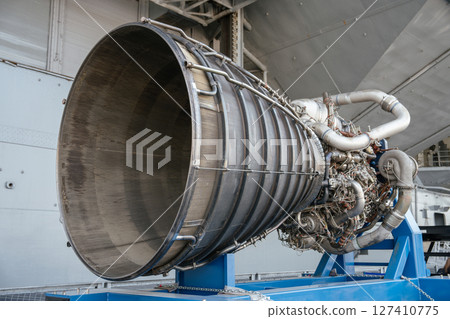 A close-up view of a powerful rocket engine with visible nozzle, pipes, and turbopump components. The engine is mounted on a blue metal frame for exhibition purposes. A close-up view of a powerful rocket engine with visible nozzle, pipes, and turbopump components. The engine is mounted on a blue metal frame for exhibition purposes. 127410775