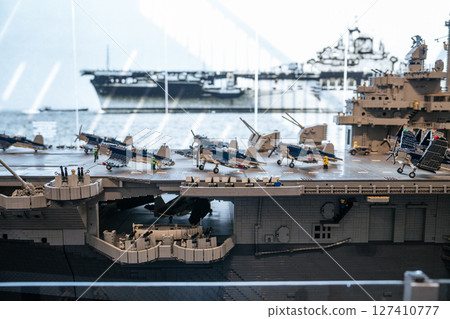 Detailed close-up of a model aircraft carrier deck lined with miniature fighter jets and crew figurines. The backdrop includes a historical photo of a real warship on the water. 127410777