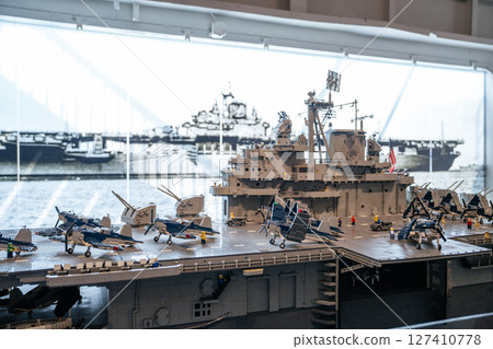 Close-up view of a model aircraft carrier featuring its command tower and miniature fighter jets on the flight deck. The background includes a historic photo of a real carrier, adding a sense of 127410778