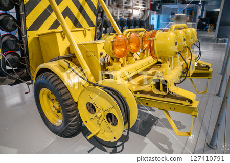 The image shows a detailed view of an airport or carrier trailer equipped with amber signal lights and thick black cables. The yellow-painted machinery is part of a guidance or signaling system 127410791