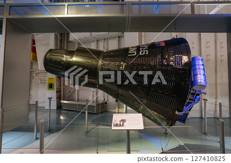 A full view of the space capsule replica exhibited in a museum. The dark ribbed surface and American flag mark its historic aerospace design. 127410825