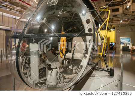 A detailed close-up of a helicopter cockpit with glass bubble canopy on display in a museum. Control levers, seats, and instrument panels are visible through the clear dome. 127410833