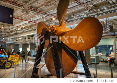 A massive bronze ship propeller is displayed upright on a metal stand inside a maritime museum. Visitors walk around the exhibit under bright industrial-style ceiling lights. A massive bronze ship propeller is displayed upright on a metal stand inside a maritime museum. Visitors walk around the exhibit under bright industrial-style ceiling lights. 127410840
