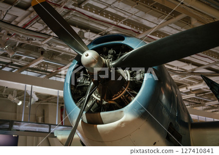 A detailed close-up of a restored vintage propeller aircraft focuses on the engine and propeller blades. The aircraft is part of an indoor aviation museum display with overhead lighting. A detailed close-up of a restored vintage propeller aircraft focuses on the engine and propeller blades. The aircraft is part of an indoor aviation museum display with overhead lighting. 127410841