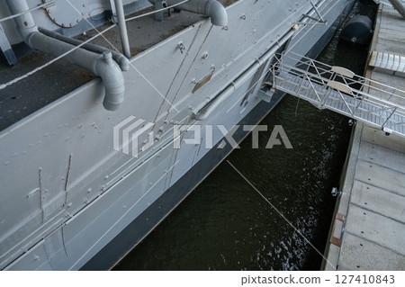 A detailed view of the gray hull of a docked aircraft carrier shows pipes, bolts, mooring ropes, and a boarding gangway. The ship is anchored alongside a concrete pier above dark water. A detailed view of the gray hull of a docked aircraft carrier shows pipes, bolts, mooring ropes, and a boarding gangway. The ship is anchored alongside a concrete pier above dark water. 127410843