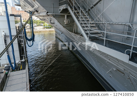 The photo captures a close-up view of an aircraft carrier's gray hull with mooring ropes and technical pipes. A staircase, handrails, and a waterline emphasize the ship's massive scale. 127410850