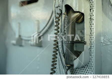 An aged black telephone handset is mounted on a metal wall aboard a military ship. The handset is secured with a zip tie and hangs next to a coiled communication cord. 127410855