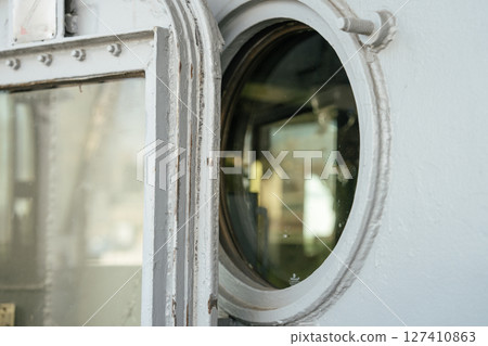 Close-up of a weathered ship door and round porthole window. Reflections of light and blurred surroundings are visible on the glass surface. 127410863
