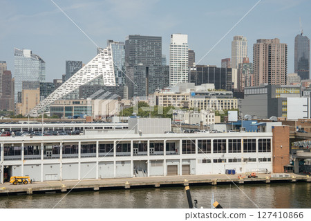 A modern triangular building stands out among New York City skyscrapers. The foreground shows a waterfront warehouse and construction vehicles near the Hudson River. A modern triangular building stands out among New York City skyscrapers. The foreground shows a waterfront warehouse and construction vehicles near the Hudson River. 127410866