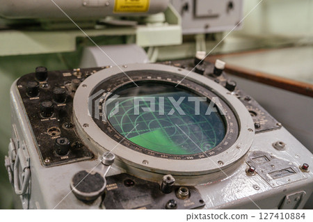 Old naval radar console with a circular green screen and mechanical controls. The radar shows a scanning grid used for detecting objects at sea. 127410884