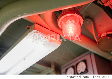 A close-up of a glowing red warning light next to a white fluorescent light on a metal ceiling. Industrial pipes and gauges are visible in the background. 127410885