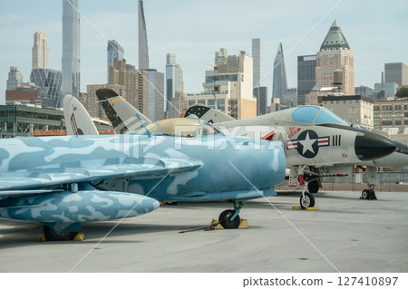 Two vintage fighter jets are showcased on an open deck against the backdrop of modern New York skyscrapers. The photo features detailed views of the aircraft fuselage and tail design. 127410897