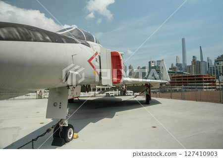 A close-up angle of a military jet fighter shows its cockpit, engine intake, and landing gear. The aircraft is parked on a rooftop with a city skyline in the background. 127410903