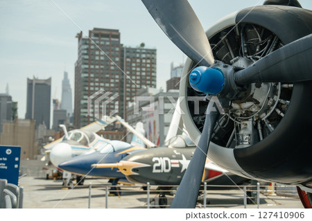A close-up of a vintage military propeller engine is displayed with other aircraft and New York City skyscrapers in the background. The black propeller blades and exposed engine parts dominate the 127410906