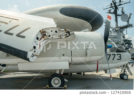 A detailed view of a white military propeller plane shows an open engine maintenance panel and exposed mechanical parts. The aircraft is parked on a tarmac near a naval radar tower. 127410908