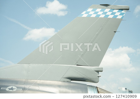 Close-up of a gray military jet tail fin featuring a blue and white checkerboard pattern near the top. The vertical stabilizer is shown against a bright sky with clouds. Close-up of a gray military jet tail fin featuring a blue and white checkerboard pattern near the top. The vertical stabilizer is shown against a bright sky with clouds. 127410909