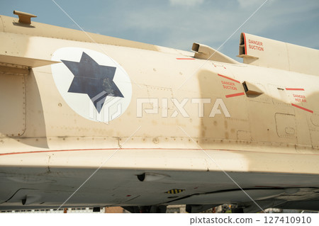 Close-up of a beige Israeli military aircraft fuselage showing a blue star emblem. Red danger warnings, suction vents, and hot air exhausts are visible on the surface. Close-up of a beige Israeli military aircraft fuselage showing a blue star emblem. Red danger warnings, suction vents, and hot air exhausts are visible on the surface. 127410910
