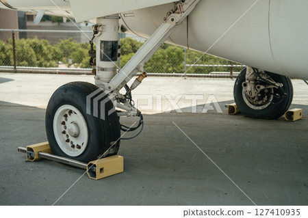Detailed view of a military aircraft's landing gear with wheels secured by chocks. The metal structure and tire textures are clearly visible on the concrete ground. Detailed view of a military aircraft's landing gear with wheels secured by chocks. The metal structure and tire textures are clearly visible on the concrete ground. 127410935