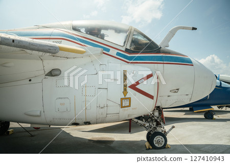 Detailed close-up of a vintage military aircraft showing the cockpit, rescue instructions, and front landing gear. The white fuselage with red and blue stripes is parked outdoors on a sunny day. 127410943