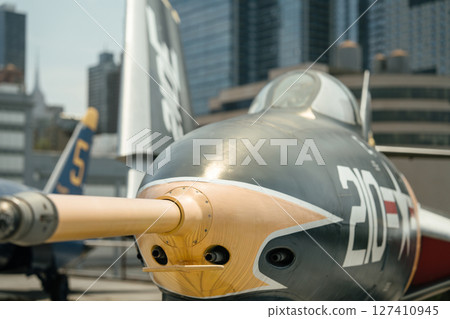 A detailed view of the nose and canopy of a vintage military jet marked with the number 210. Gun ports and a painted nose cone are clearly visible with urban buildings in the background. A detailed view of the nose and canopy of a vintage military jet marked with the number 210. Gun ports and a painted nose cone are clearly visible with urban buildings in the background. 127410945