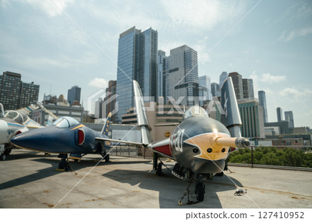 Two vintage U.S. Navy jet fighters are showcased on an aircraft carrier deck with a modern city skyline behind. The military planes are preserved for public exhibition as part of an aviation museum. 127410952