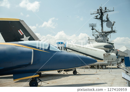 Close-up of the nose and cockpit of Navy jet painted in blue and yellow. The aircraft is stationed on a carrier deck with radar structures and other military planes in the background. 127410959