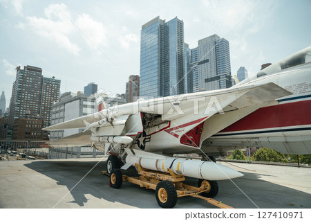 White and red military jet aircraft with a mounted missile shown from the side on a museum deck. Tall skyscrapers of New York City rise in the background under a sunny sky. 127410971