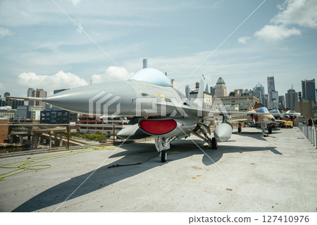 Wide view of a fighter jet with red intake cover and other aircraft lined up on an outdoor museum deck. The background features the skyline of New York City under a sunny sky. Wide view of a fighter jet with red intake cover and other aircraft lined up on an outdoor museum deck. The background features the skyline of New York City under a sunny sky. 127410976
