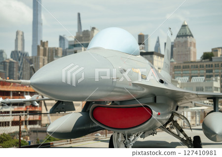 Detailed close-up of jet nose, cockpit, and red intake cover on display. The aircraft is parked outdoors at an aviation museum with a city skyline behind. Detailed close-up of jet nose, cockpit, and red intake cover on display. The aircraft is parked outdoors at an aviation museum with a city skyline behind. 127410981