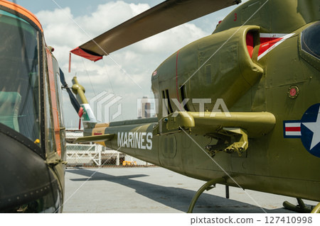 Back side of a green military helicopter labeled MARINES on an outdoor museum deck. The rotor blade, tail, and surrounding aircraft are visible with city buildings in the background. Back side of a green military helicopter labeled MARINES on an outdoor museum deck. The rotor blade, tail, and surrounding aircraft are visible with city buildings in the background. 127410998