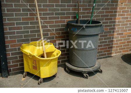 A yellow mop bucket with a wooden handle and a large gray trash bin with wheels stand beside a brick wall. Cleaning tools are positioned upright under bright outdoor sunlight. 127411007