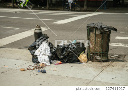 Black garbage bags and loose trash lie next to an overflowing street bin on a concrete sidewalk. A fire hydrant and urban crosswalk mark the busy city setting. 127411017