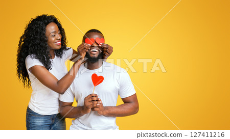 Black young woman surprising her beloved man with valentine gift, closing his eyes with red love cards, yellow studio background 127411216